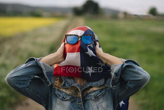 Signora in abiti casual soggiorno in campo giallo con occhiali da sole sopra la bandiera americana con la luce del sole — Foto stock