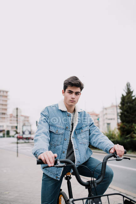 Jovem adolescente bonito inclinado no guidão da bicicleta e olhando para a câmera na rua — Fotografia de Stock