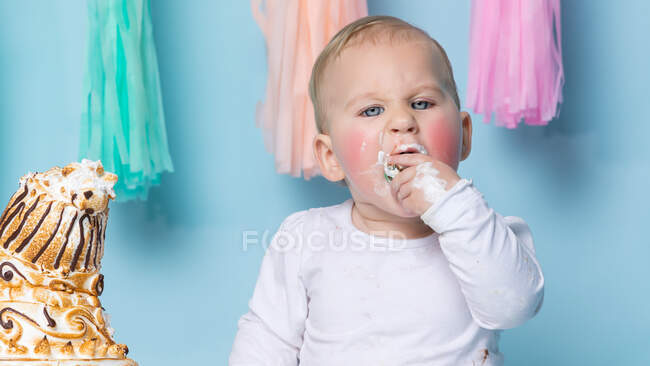 Mignon petit garçon assis au gâteau d'anniversaire et le manger avec la main sur fond bleu. — Photo de stock