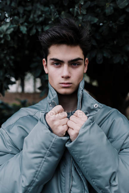 Junger hübscher Teenager in grauer schicker warmer Jacke steht vor grünem Baum — Stockfoto