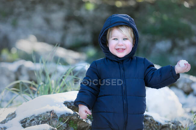 Cheerful little toddler boy looking at camera and holding snowball in nature. — Stock Photo