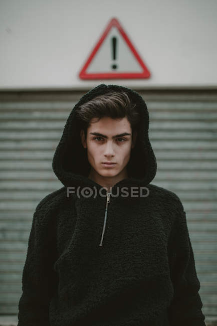 Thoughtful teenager in black hooded jacket standing on street with exclamation sign and looking at camera — Stock Photo
