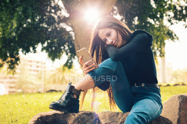 Jeune femme souriante assise sur le rocher et utilisant un smartphone dans le parc — Photo de stock