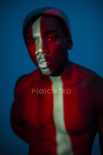 Безэмоциональный этнический мужчина, стоящий со световой линией на теле и смотрящий в камеру — стоковое фото