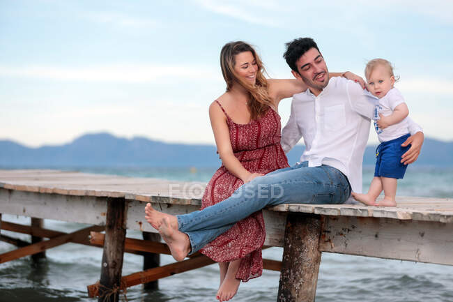 Feliz madre y padre sentados con el niño en el muelle de madera. - foto de stock