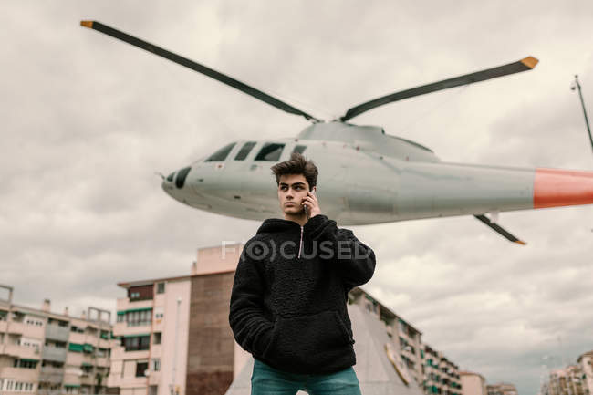 Красивий молодий чоловік стоїть біля пам'ятника вертольоту в місті і розмовляє по смартфонах — стокове фото
