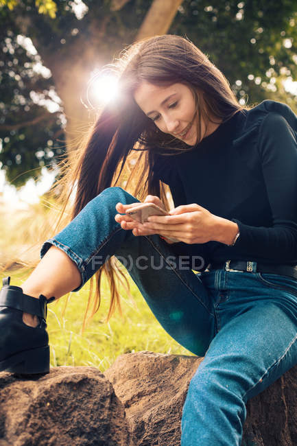 Jovem mulher sorridente sentada no rock e usando smartphone no parque — Fotografia de Stock