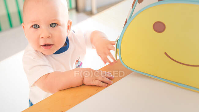 Очаровательный маленький мальчик сидит у красочной сумки с улыбкой на лице и смотрит в камеру. — стоковое фото