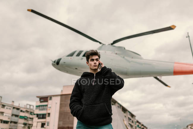 Bello giovane uomo in piedi al monumento elicottero in città e parlando su smartphone — Foto stock