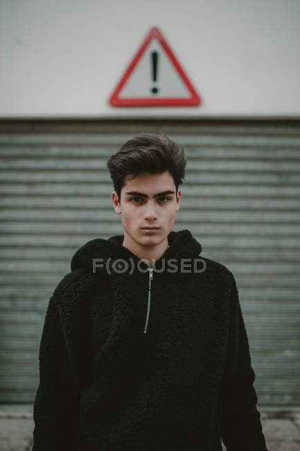 Pensativo adolescente en negro chaqueta con capucha de pie en la calle con signo de exclamación y mirando a la cámara - foto de stock