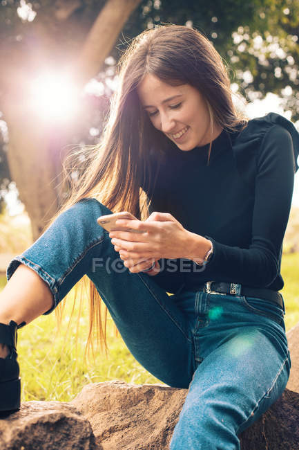 Jovem mulher sorridente sentada no rock e usando smartphone no parque — Fotografia de Stock
