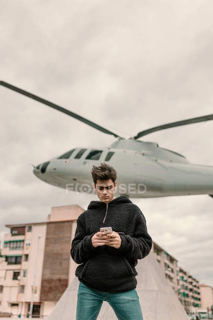 Bello giovane uomo in piedi al monumento elicottero in città e utilizzando smartphone — Foto stock