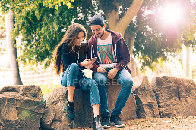Rindo jovem casal sentado no rock com smartphone no parque — Fotografia de Stock