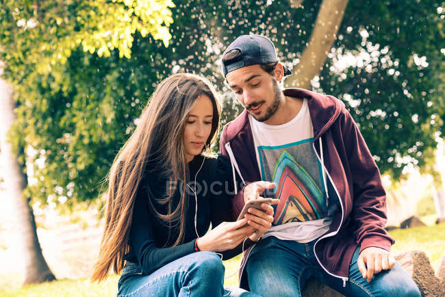 Junges Paar sitzt im Park und teilt Smartphone — Stockfoto
