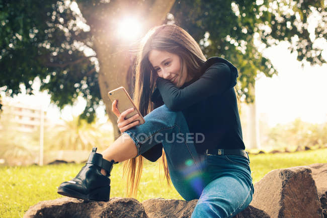 Jeune femme souriante assise sur le rocher et utilisant un smartphone dans le parc — Photo de stock