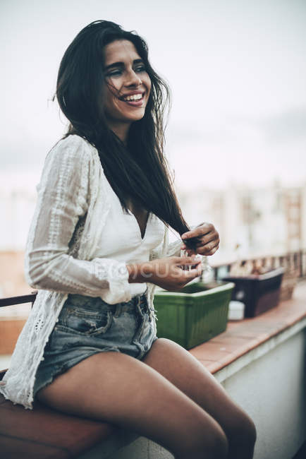 Молодая смеющаяся женщина в рубашке и джинсовых шортах сидит на балконе — стоковое фото