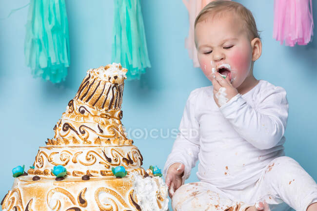 Lindo niño sentado en el pastel de cumpleaños y comerlo con la mano sobre fondo azul. - foto de stock