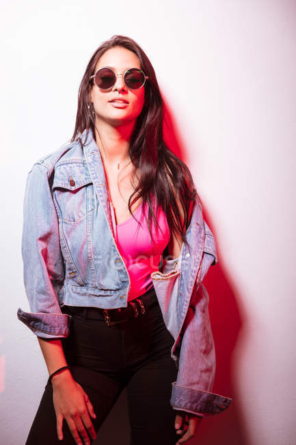 Bella giovane donna vestita di rosa con gli occhiali da sole in piedi al muro bianco e guardando la fotocamera — Foto stock
