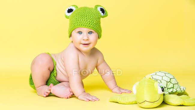 Entzückender kleiner Junge mit Froschhut sitzt auf Schildkrötenspielzeug auf gelbem Hintergrund. — Stockfoto
