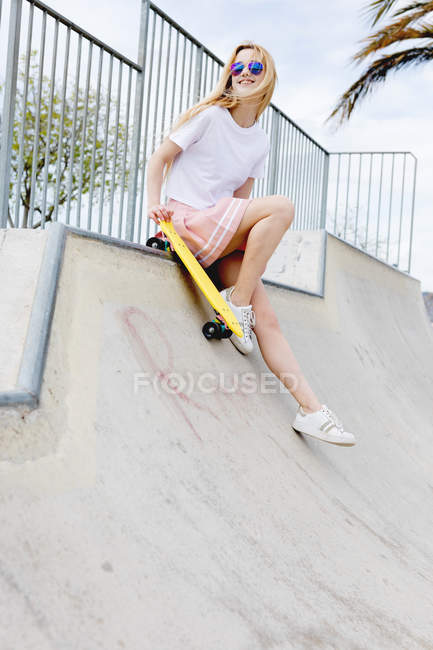 Sonriente chica rubia con estilo en gafas de sol con penny board en skate park - foto de stock