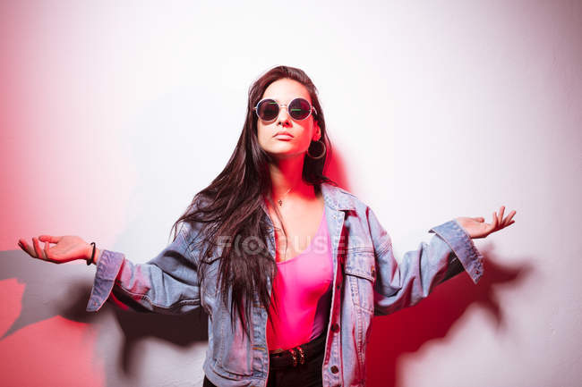 Pretty young pink dressed woman in sunglasses standing at white wall with arms outstretched — Stock Photo