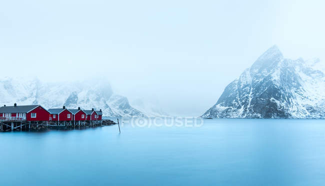 Ландшафт маленьких красных деревянных домиков на побережье против снежных гор в тумане, Норвегия — стоковое фото