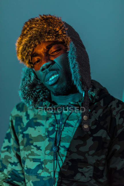 Hombre negro despectivo en ropa de abrigo y sombrero con color naranja en la cara - foto de stock