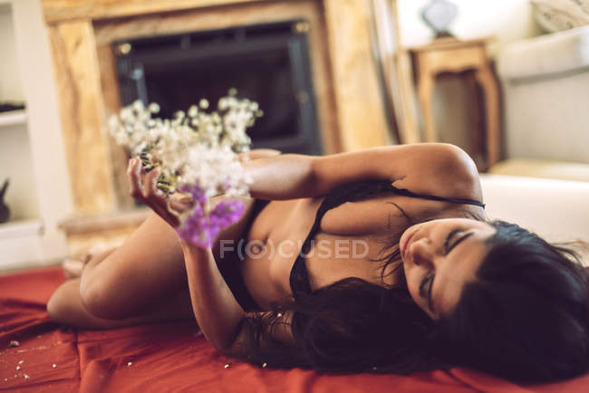 Joven mujer seductora acostada en sábana roja en lencería y sosteniendo flores frescas - foto de stock
