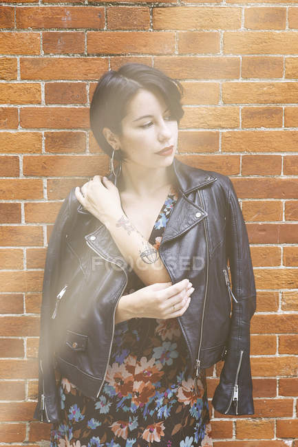 Thoughtful tattooed woman in patterned dress and leather jacket standing in front of brick wall — Stock Photo