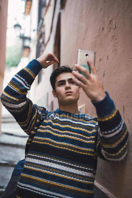 Adolescente seguro de sí mismo en suéter de pie en la calle de la ciudad vieja y tomar selfie con teléfono inteligente - foto de stock