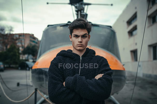 Young doubtful man standing with arms crossed with helicopter monument on background — Stock Photo