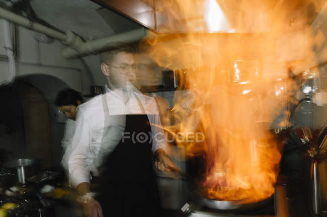 Cocinero haciendo un flambe en la cocina del restaurante con colega en el fondo - foto de stock