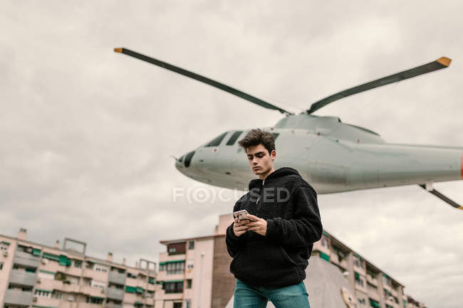 Bonito jovem de pé no monumento de helicóptero na cidade e usando smartphone — Fotografia de Stock