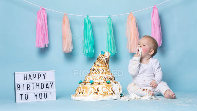 Cute little boy sitting at birthday cake and eating it with hand on blue background. — Stock Photo