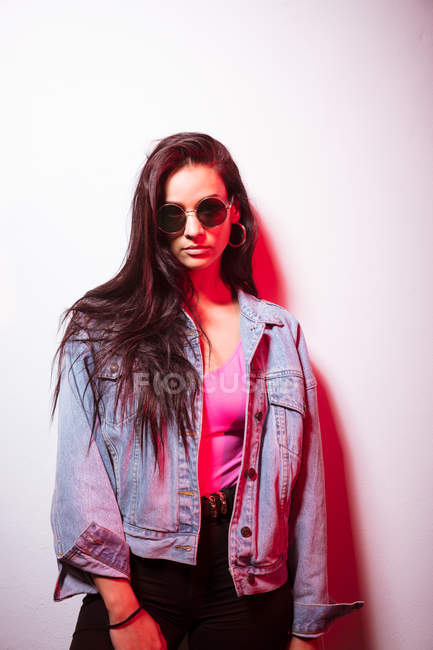 Muito jovem rosa vestido mulher em óculos de sol em pé na parede branca e olhando para a câmera — Fotografia de Stock