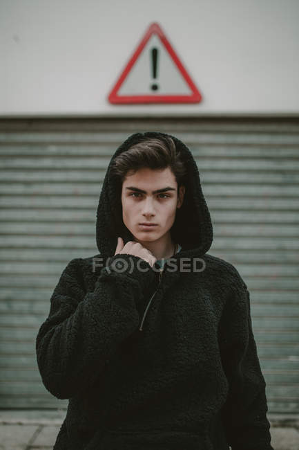 Pensativo adolescente en negro chaqueta con capucha de pie en la calle con signo de exclamación y mirando a la cámara - foto de stock