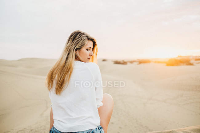 Jeune femme en t-shirt blanc assis sur le sable au coucher du soleil et regardant par-dessus l'épaule — Photo de stock