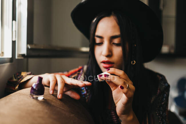 Досить молода жінка в стильному вбранні, використовуючи пензлик для нанесення лаку для нігтів, сидячи на зручному дивані вдома — стокове фото