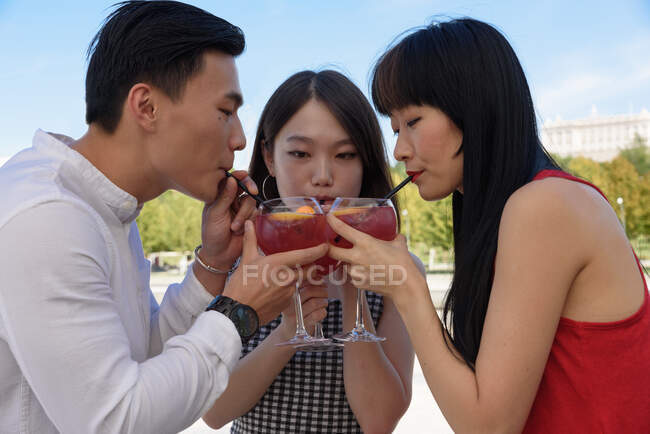 Three Asian people clinking glasses and using straws to enjoy delicious cold drink while standing on city street on sunny day — Stock Photo