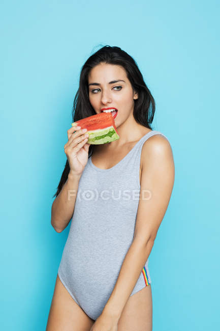 Чувственная брюнетка в сером телесном костюме ест свежий арбуз и смотрит в сторону на голубом фоне — стоковое фото