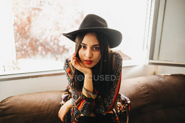 Attractive young lady in stylish outfit smiling and keeping eyes closed while sitting on back of soft sofa near window — Stock Photo