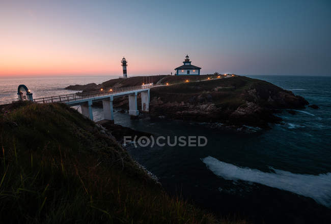 Modern illuminated bridge located near small islet with beacon and house at dusk, Asturias, Spain — Stock Photo