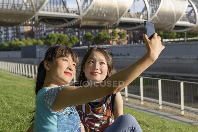 Zwei reizende asiatische Damen lächeln und posieren für ein Selfie, während sie an einem sonnigen Tag im Park auf dem Boden sitzen — Stockfoto