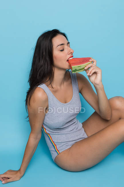 Young sensual woman in bodysuit holding piece of watermelon on blue background — Stock Photo