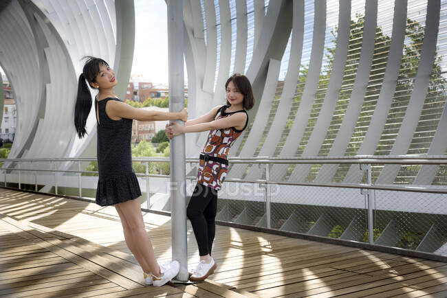 Side view of pretty Asian ladies smiling and looking at camera while standing near metal pole on modern bridge in park — Stock Photo