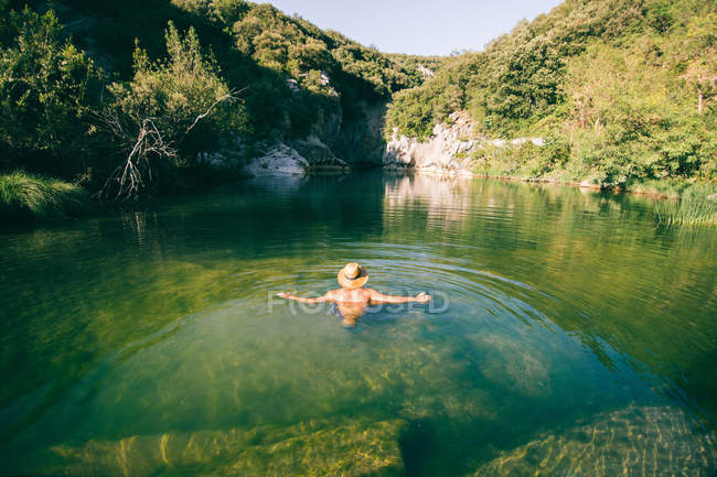 Rückansicht des hemdlosen Typen mit Hut, der im sauberen Wasser eines erstaunlichen Sees in den Bergen steht — Stockfoto