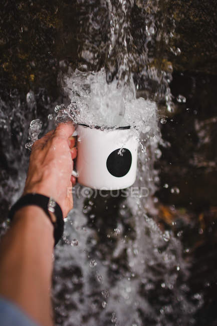 Mano del hombre sosteniendo jarra bajo agua dulce de fuente de agua fría en la naturaleza - foto de stock