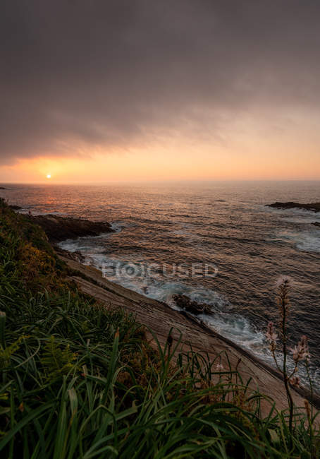 Stormy sea splashing near slope of grassy coast at sunset in cloudy evening, Asturias, Spain — Stock Photo