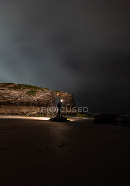Силуэт анонимного человека с ярким фонариком, стоящего на берегу у моря и скалы в пасмурную ночь, Астурия, Испания — стоковое фото