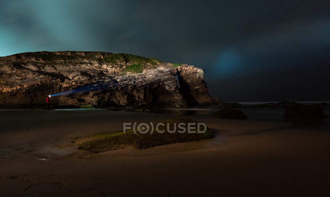 Silhueta de pessoa anônima com lanterna brilhante em pé na costa perto do mar e penhasco na noite nublada, Astúrias, Espanha — Fotografia de Stock